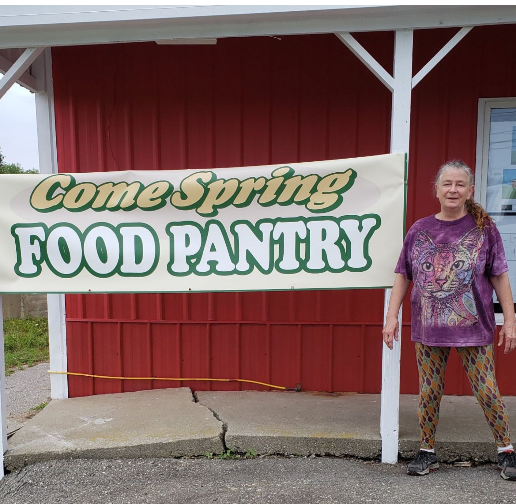 Tracy standing in front of the red come spring food pantry building in union maine with a new sign that says come spring food pantry in bubble letters for the grand opening.