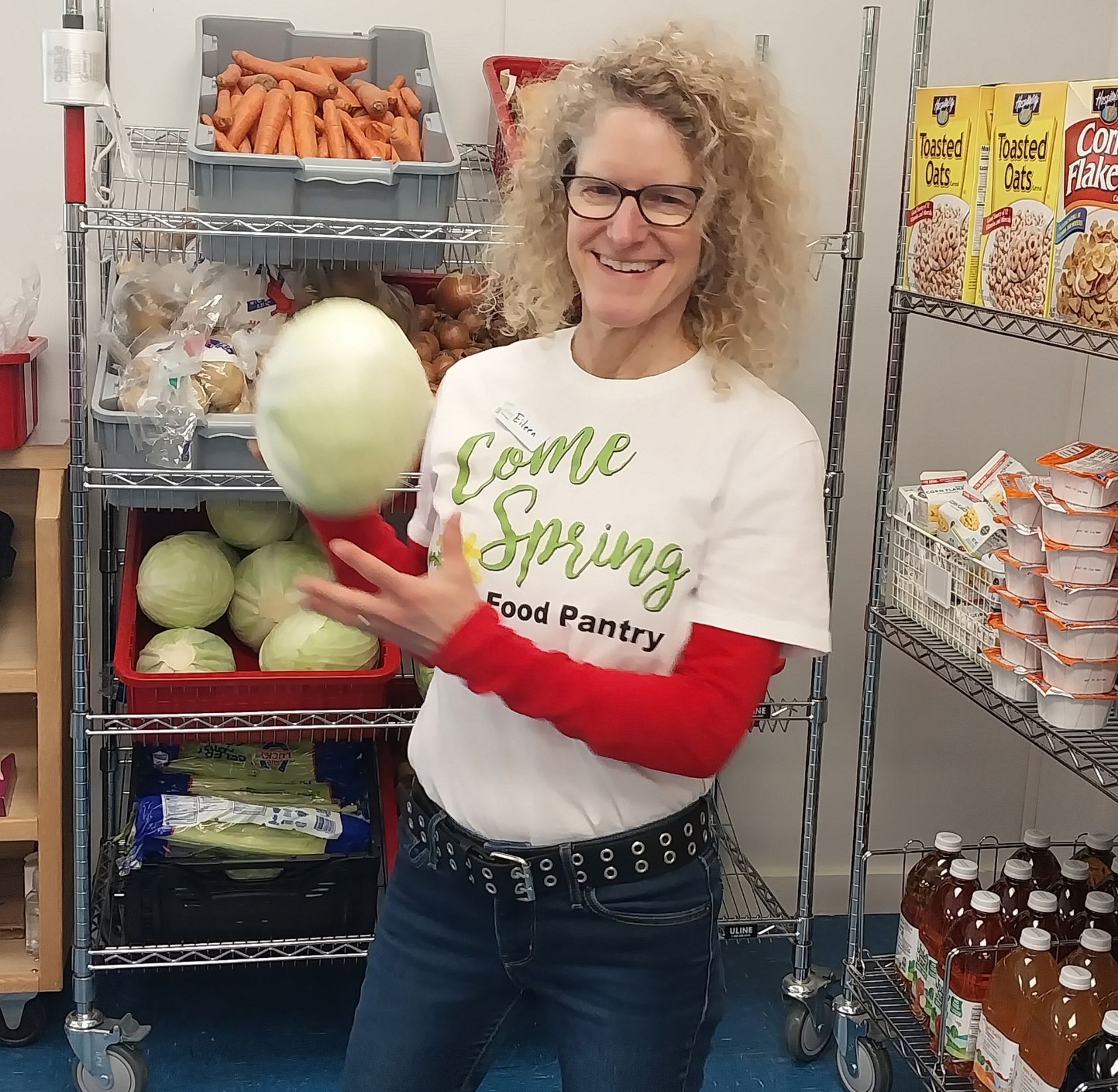 woman in come spring food pantry branded shirt and red sleeves tossing a cabbage and smiling
